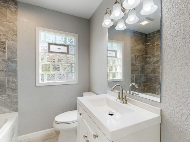 A modern bathroom featuring a white vanity, mirror, and tiled wall lit by natural light.
